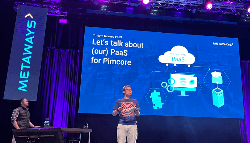 Two men on a stage, in the background a screen with the title of the keynote "Let's talk about (our) PaaS for Pimcore"